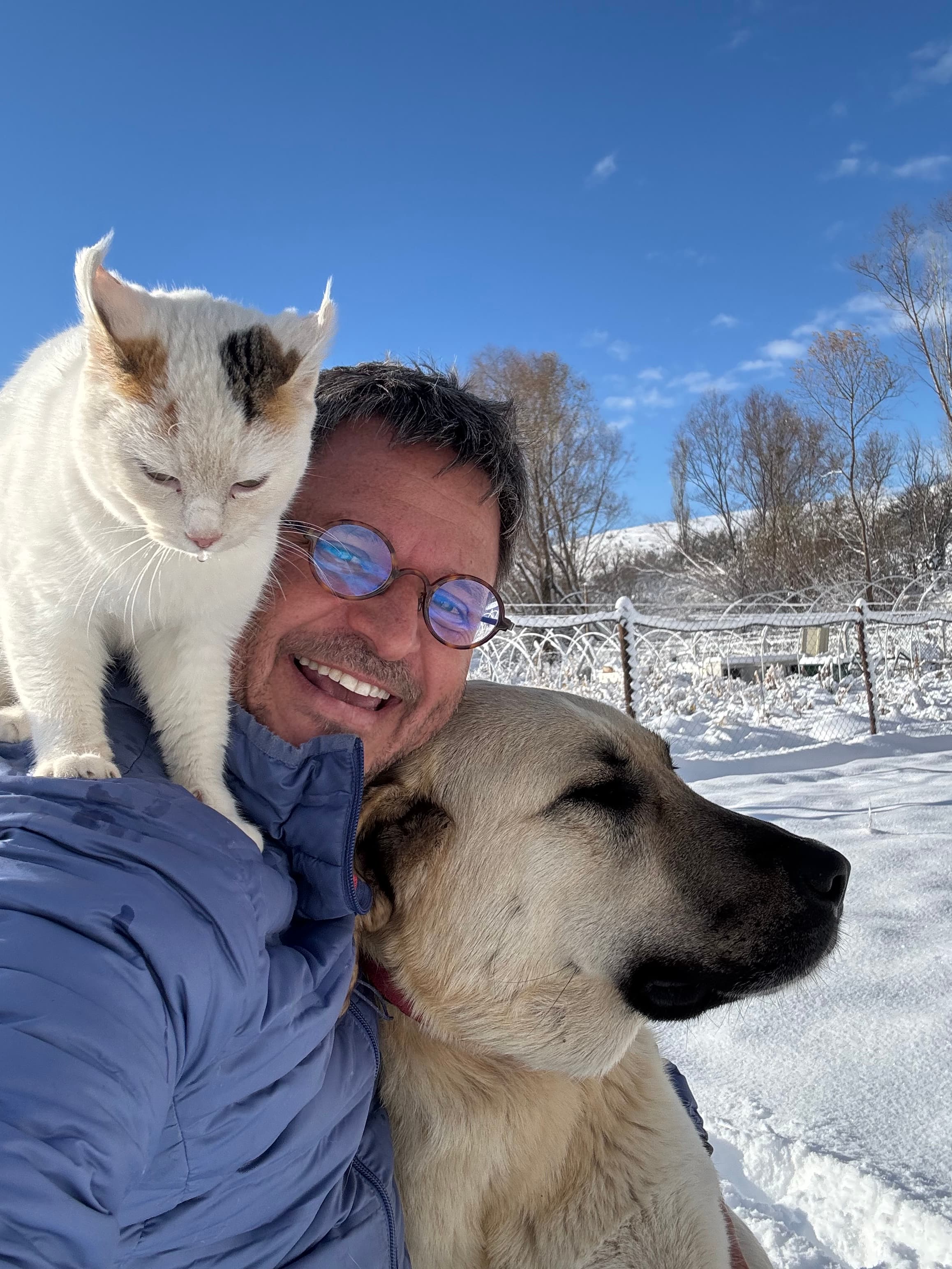 Smiling man with glasses holding a white cat and large dog in a snowy landscape.